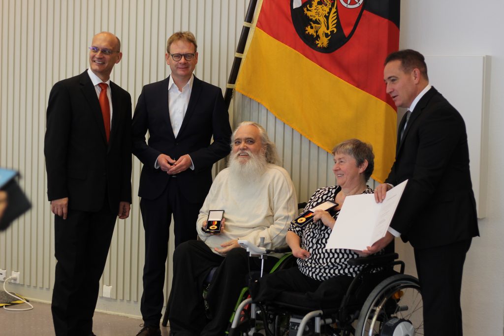 Erika Naumer-Klein und Robert Schneider sitzend im Rollstuhl mit ihren Verdienstorden in der Hand. Dahinter die Laudatoren und die Landesflagge von Rheinland-Pfalz.