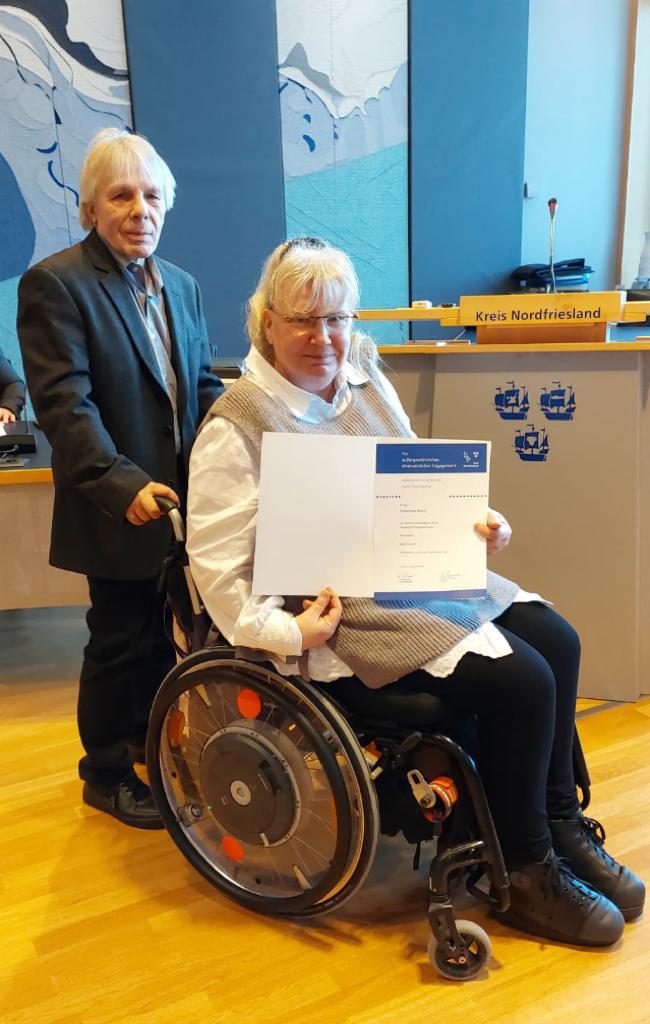 Rosemarie Böing präsentiert im Rollstuhl sitzend ihre Urkunde